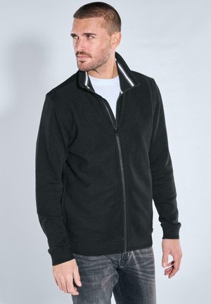Schwarze Fleece-zip-up-Jacke mit Stehkragen, gerippten Akzenten und langen Ärmeln, kombiniert mit einem weißen T-Shirt und grauen Jeans.