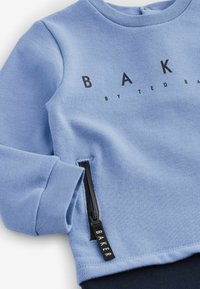 Světlemodrá mikina s černým nápisem "BAKER"; obsahuje boční zipovou kapsu s černým zipem a strukturovanou tkaninou. Kulatý výstřih a manžety na rukávech.