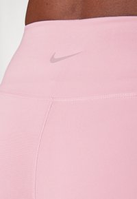 Ružičaste legice visoko pasu z gladko teksturo, ki vsebujejo subtilen sivi Nike logotip blizu pasu in kontrastne šive.
