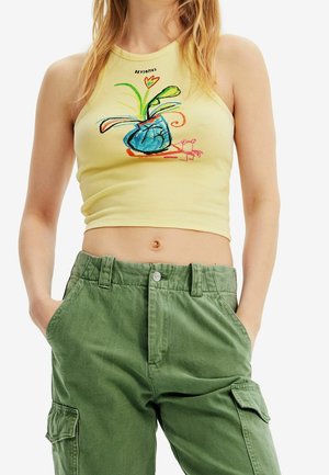 Frau trägt ein hellgelbes, kürzer geschnittenes Tanktop mit einem bunten abstrakten Pflanzendesign und hoch taillierte grüne Cargo-Hosen, die Hände in den Taschen.