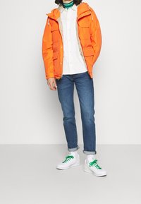 Person med en orange jacka över en vit skjorta, blå uppvikkade jeans och vita sneakers med gröna snören, stående mot en enkel bakgrund.