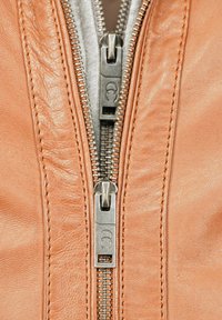 Braune Lederjacke mit einem zentralen silbernen Metallreißverschluss und doppeltem Zug, umrandet von feinen Nähten auf beiden Seiten.