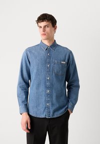 Lichtblauw denim overhemd met knopen, lange mouwen, een kraag en een borstzak. Voorzien van witte knopen en zichtbare stikseldetails.