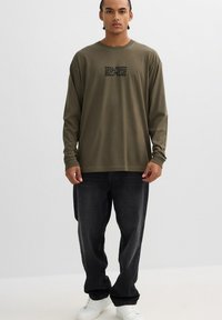 Camisa de manga larga de color oliva con texto negro que dice "CÁE SIETE VECES PERO LEVÁNTATE OCHO VECES"; combinada con jeans oscuros y zapatillas blancas.