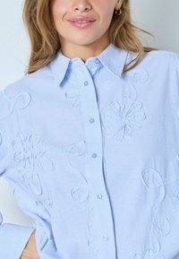 Vaaleansininen raidallinen napillinen paita, jossa kohokuvioitu kukkakirjailu ja hienovaraiset kiemuraiset kuviot. Lyhyet hihat ja klassinen kaulussuunnittelu.