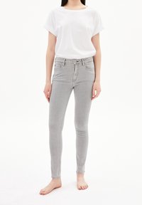 Jeans skinny de color gris claro con cintura alta, cinco bolsillos y cierre de botón, combinados con una camiseta blanca de corte holgado.