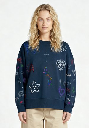 Donna con capelli biondi ondulati indossa una felpa blu navy con scarabocchi colorati, simboli e testo, abbinata a pantaloni beige su sfondo semplice.