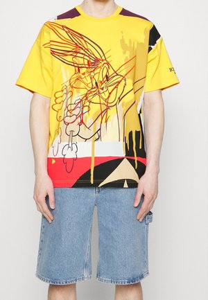 Person trägt ein gelbes abstraktes Bugs Bunny Grafik-T-Shirt und hellblaue Jeansshorts und steht vor einem neutralen Hintergrund.