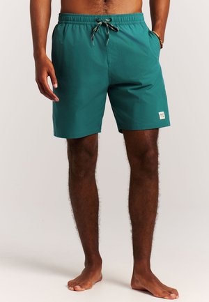 Man draagt turquoise zwemshorts met een trekkoord in de taille en zijzakken, staat blootsvoets tegen een effen achtergrond.