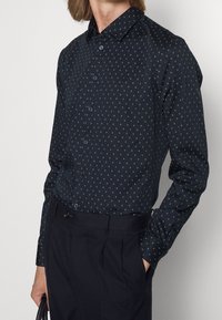 Tmavě modrá košile s dlouhými rukávy a geometrickým vzorem malých zlatých diamantes. Má límec na knoflíky a modré knoflíky.