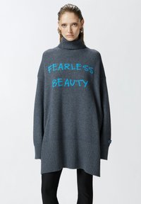 Prevelik sivi pulover s visokim ovratnikom. Sadrži plavi natpis „BEZ STRAHA LJEPOTA“ u debelim slovima. Rebrasti rub i manžeti, mekana tekstura.