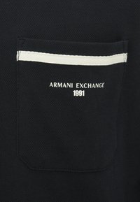 Svart plagg med teksturert stoff som har en lommepatch. Hvit dekorativ stripe på lommen. Merket leser "ARMANI EXCHANGE" og "1991."