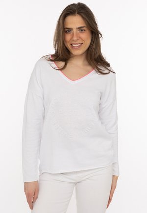 Junge Frau mit langen braunen Haaren, die ein weißes Langarmshirt mit V-Ausschnitt und weiße Hosen trägt, lächelt und steht vor einem weißen Hintergrund.