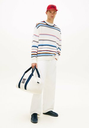 Junger Mann in einem mehrfarbigen, gestreiften Pullover, weißen Hosen, roter Mütze, schwarzen Sneakers, der eine weiße Tommy Jeans Reisetasche an den Griffen hält.