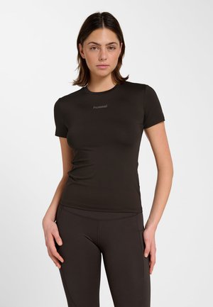 Frau trägt eng anliegendes schwarzes Sportshirt mit kurzen Ärmeln und passende Leggings, steht mit den Händen auf den Oberschenkeln vor schlichtem weißem Hintergrund.