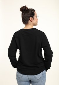 Vrouw met donker haar in een knot, die een bril draagt, een zwarte lange mouwen shirt en lichte spijkerbroek, gezien van achteren tegen een effen achtergrond.