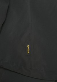 Veste en tissu noir avec une texture lisse et des détails de couture, dotée d'une étiquette jaune « TEXAPORE » en bas à gauche.