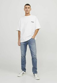 Camiseta blanca de gran tamaño con logo negro, pantalones vaqueros desgastados en azul claro y zapatillas negras y blancas con acentos a rayas.