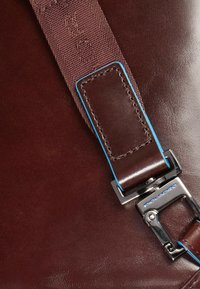 Bolso de cuero marrón con acabado brillante, con una correa de tela con detalles azules y una hebilla de metal plateado. Textura suave con detalles mínimos.