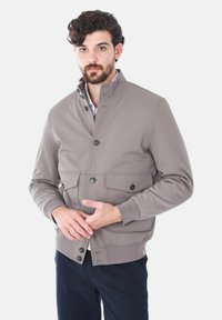 Giacca bomber grigia chiara con alto colletto, bottoni frontali, due tasche sul petto e polsini a coste. Texture liscia, design casual e forma aderente.