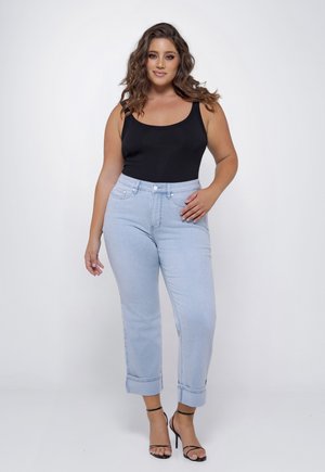 Vrouw met golvend haar, gekleed in een zwart mouwloos topje, lichtblauwe opgerolde jeans en zwarte sandalen met hoge hakken, staand tegen een witte achtergrond.