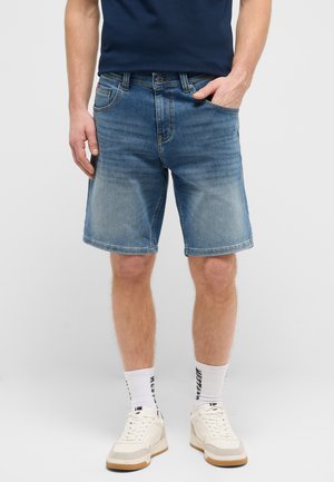 Man wearing blue denim shorts, white socks with black text, white sneakers, and a dark blue t-shirt, standing against a plain background.