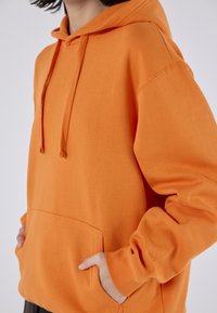 Felpa arancione con cappuccio, dotata di tasca a marsupio, texture morbida e cappuccio con cordino. Presenta una vestibilità ampia e polsini a coste.