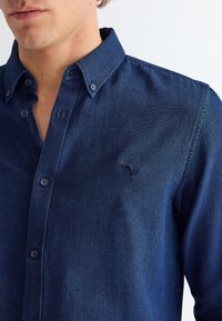 Primo piano di un uomo che indossa una camicia di jeans blu scuro con bottoni, colletto con bottoni e un piccolo logo ricamato sul petto.