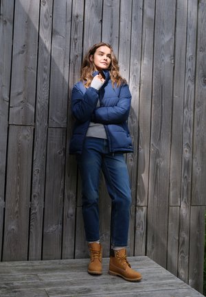 Jeune femme en veste bleue et jean, debout, bras croisés contre une haute clôture en bois, portant des bottes lacées couleur beige.