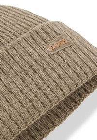 Beige gebreide beanie met een ribbeltextuur, voorzien van een rechthoekig logo-opdruk in oranje met de tekst "BOSS". Zacht, geweven stof.