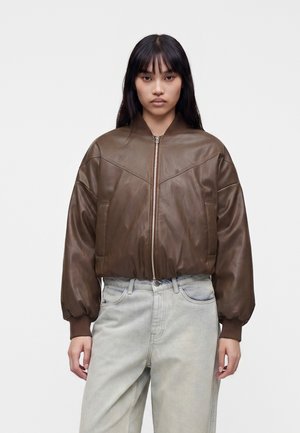 Femme aux cheveux raides et foncés portant une veste bomber en cuir marron et un jean taille haute gris clair, debout devant un fond blanc.