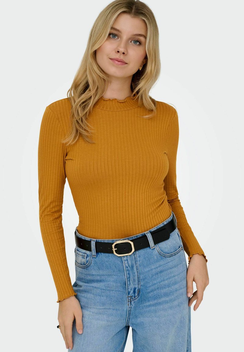 Geribbelde langwerpige top in mosterdgeel met een gekartelde halslijn, gecombineerd met lichtblauwe high-waisted jeans en een zwarte riem.