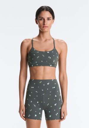 Žena stojí, oblečená v zhodný tmavozelený športový podprsenka s kvetinovým vzorom a krátke nohavice s vysokým pásom na jednoduchom bielom pozadí.