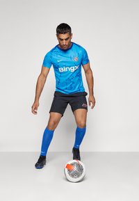 Camiseta de fútbol azul con detalles en negro, que presenta un logo y texto. Pantalones cortos negros, calcetas azules y un balón de fútbol con patrón sobre un fondo plano.