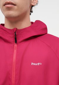 Gros plan d'une personne portant une veste zippée rose vif avec capuche et un logo blanc « huf » sur la poitrine, sur fond uni.