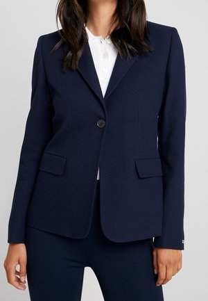 Femme portant un blazer bleu marine texturé avec un bouton sur une chemise blanche à col, debout devant un fond clair uni.