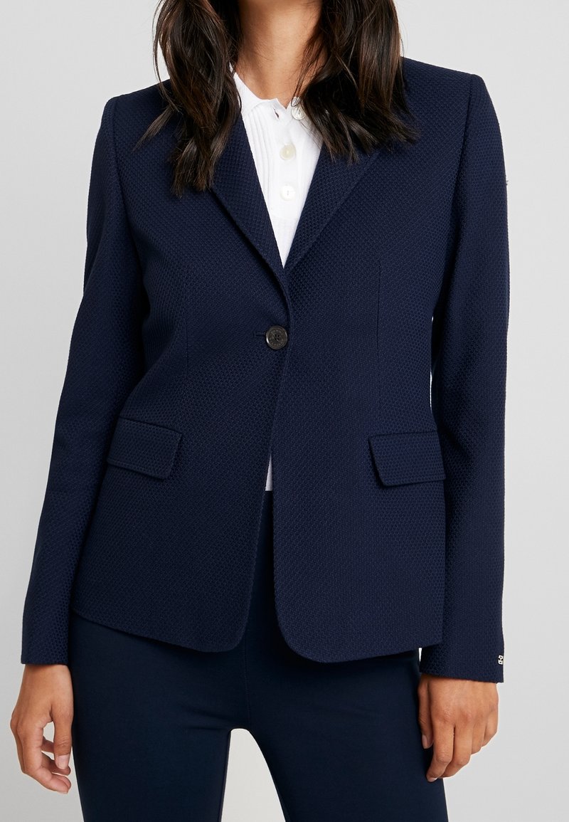 Femme portant un blazer bleu marine texturé avec un bouton sur une chemise blanche à col, debout devant un fond clair uni.
