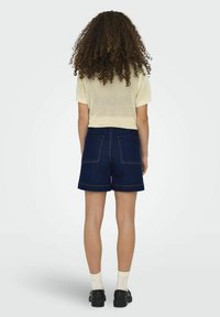 Top de punto color crema con mangas cortas y bordes en forma de onda, combinado con pantalones cortos de mezclilla oscura. Cuenta con bolsillos traseros y un corte recto. Zapatos negros y calcetines blancos.