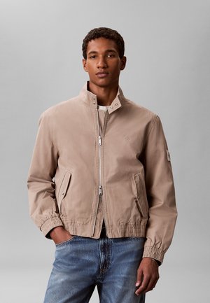 Young man wearing a beige zip-up jacket with elastic cuffs and blue jeans, standing with one hand in pocket against a plain gray background.