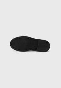 Suela de zapato negra con goma texturizada y ranuras horizontales para mayor tracción. Incluye un detalle de logotipo rectangular cerca del talón.