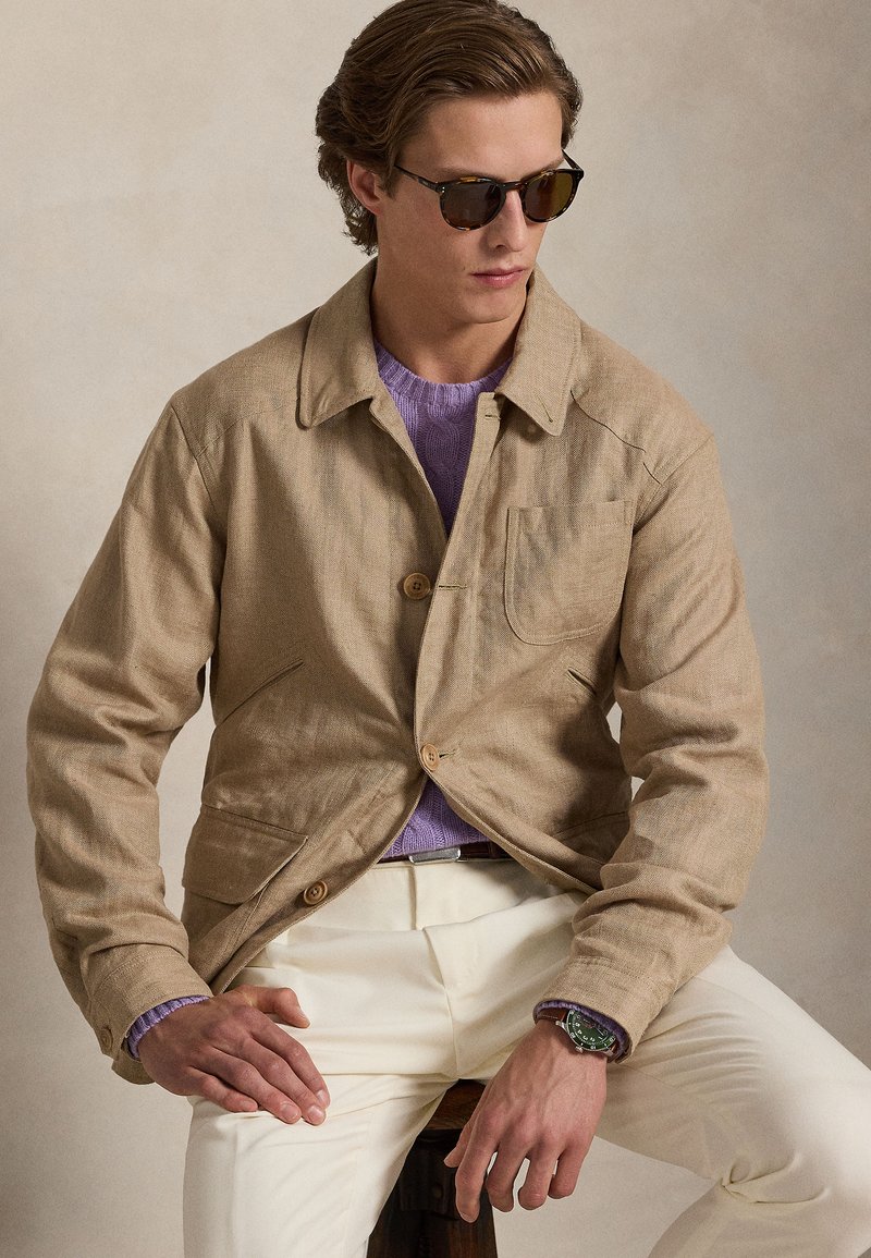 Man wearing beige jacket, lavender sweater, off-white pants, sunglasses, and green wristwatch, seated on a wooden stool against neutral background.