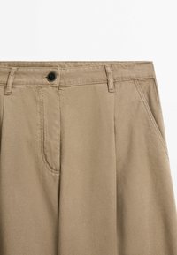 Beżowe spodnie o prostym kroju, z pojedynczym czarnym guzikiem, przednimi kieszeniami oraz gładką, solidną fakturą materiału.