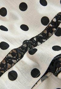 Tissu blanc à gros pois noirs avec une bordure centrale en dentelle noire ornée de motifs géométriques brodés.