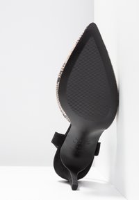 Chaussure à talon haut noire à bout pointu avec semelle texturée et bords extérieurs à motif peau de serpent, positionnée à la verticale contre des murs blancs.