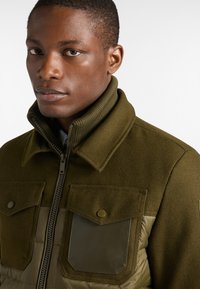 Giacca verde in misto materiale con parte superiore in lana, parte inferiore imbottita, due tasche sul petto, chiusura con zip e colletto alto. La texture appare morbida e strutturata.