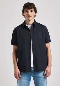 Camisa de boton corto de manga en color azul marino oscuro con acabado texturizado y un solo bolsillo en el pecho, usada sobre una camiseta blanca, combinada con jeans de color azul claro.