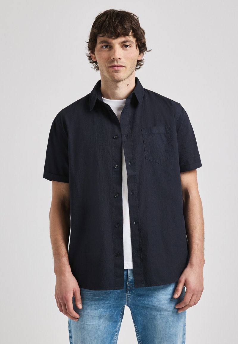 Camisa de boton corto de manga en color azul marino oscuro con acabado texturizado y un solo bolsillo en el pecho, usada sobre una camiseta blanca, combinada con jeans de color azul claro.