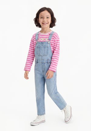 Enfant souriant aux cheveux bouclés porte un t-shirt manches longues à rayures roses, une salopette en jean et des baskets blanches montantes, debout sur une jambe.