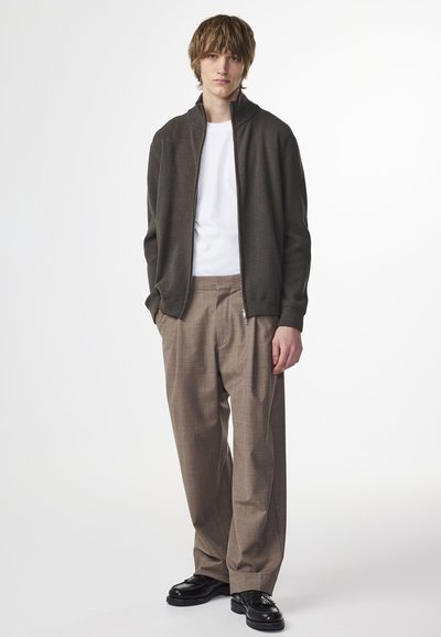 Veste bomber texturée vert foncé, t-shirt blanc, pantalon à carreaux beiges, et chaussures noires. L'ensemble est décontracté avec une coupe ample.