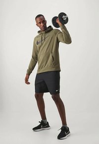 Olive rohelise kapuutsiga särk musta Nike logo ja mustade lühikeste pükste ning mustade spordijalatsitega, hoides ühes käes hantlit.